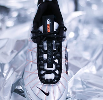A single sneaker with a sleek, modern design lies on a metallic, crumpled background. The shoe features a white base with black laces and accents, as well as orange stitching detail. The brand's name is visible on the tongue of the shoe.