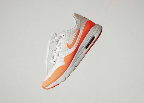 A stylish athletic sneaker featuring a white and orange color scheme, with perforated detailing and a visible air cushion in the sole. The shoe is positioned against a plain background, emphasizing its sleek and modern design.
