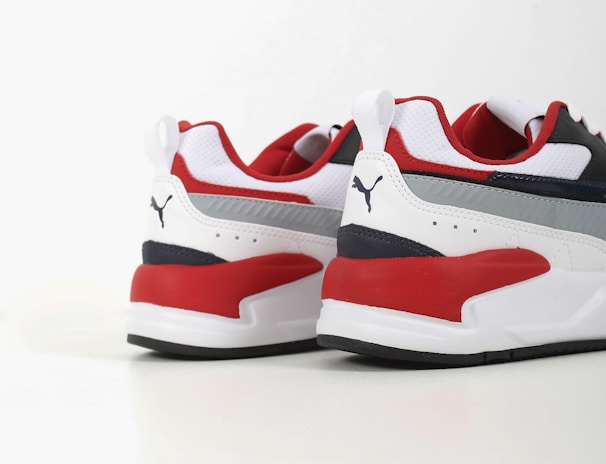 Two athletic shoes with bold red, white, and dark blue color blocking. The shoes feature a sleek, modern design with a flat sole and a recognizable logo in black on the heel. The upper part is made of a mix of textiles with some parts having perforations for breathability.